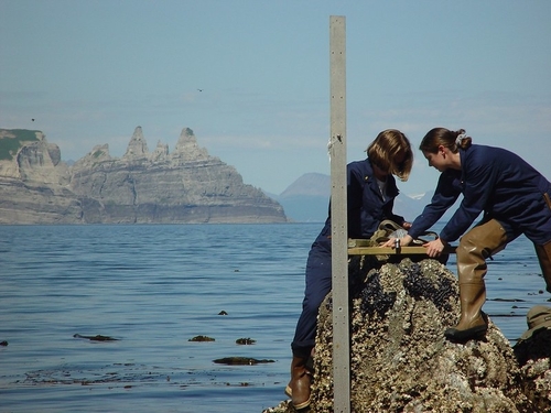 Two women wearing rubber boots, standing on rocks at the shore and installing a tide gage instrument.
