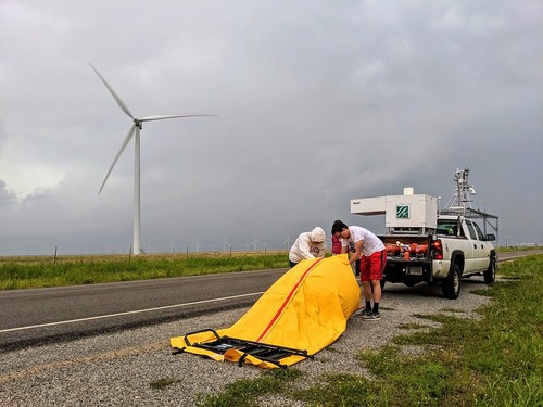 Two technicians removing gear from a large yellow bag.  Pickup truck with a variety of weather instruments mounted on top.  A wind turbine and grey clouds in the background.